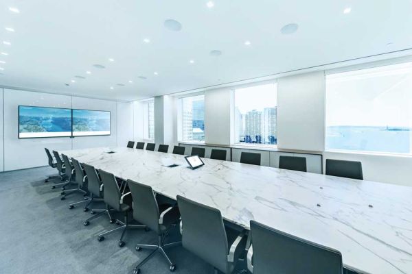 A bright executive boardroom with a long, white marble conference table, surrounded by sleek gray office chairs. Large windows offer a city skyline view in the background.