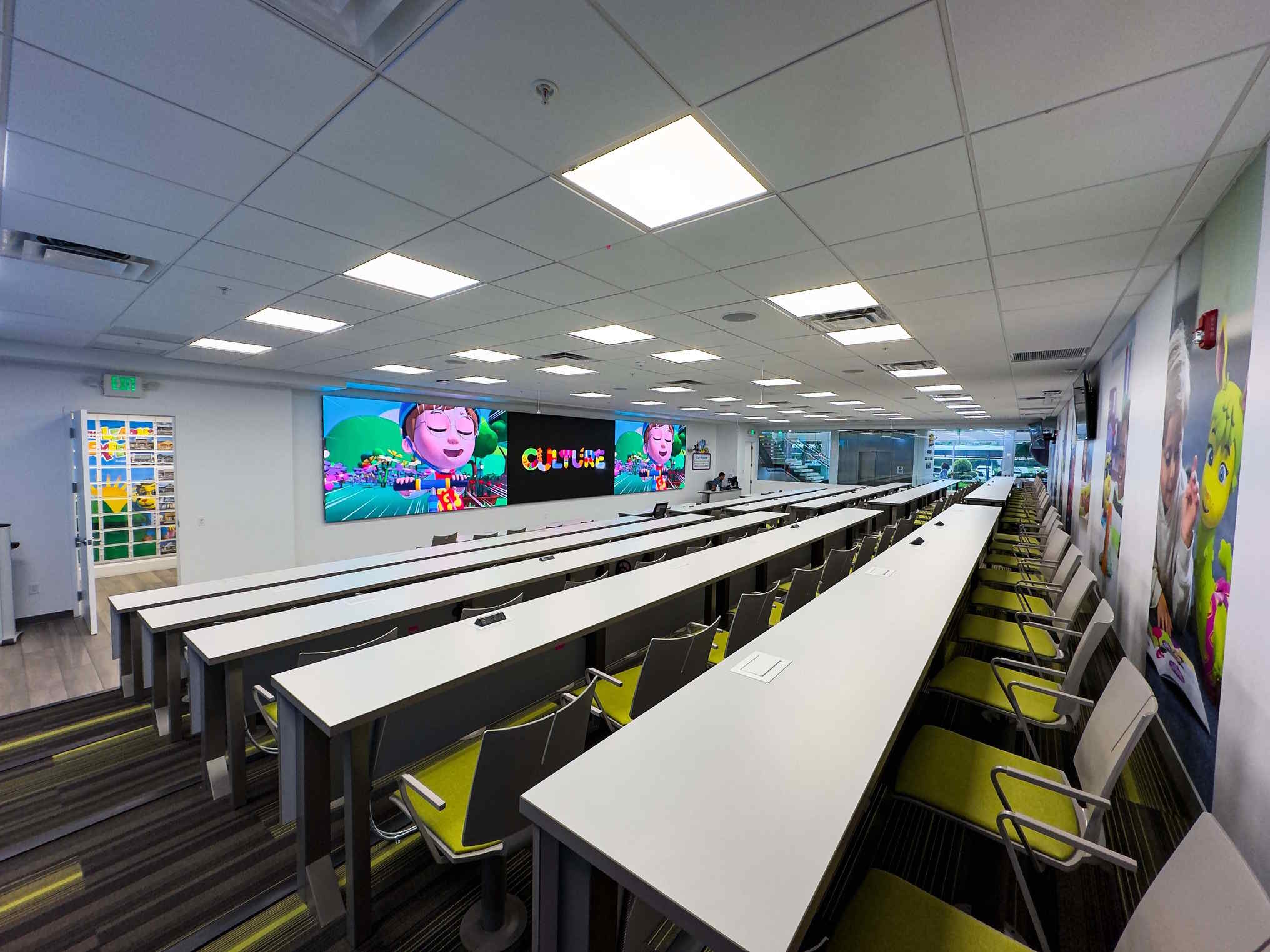 Bright classroom-style space with white tables, green chairs, and large digital screens at the front.