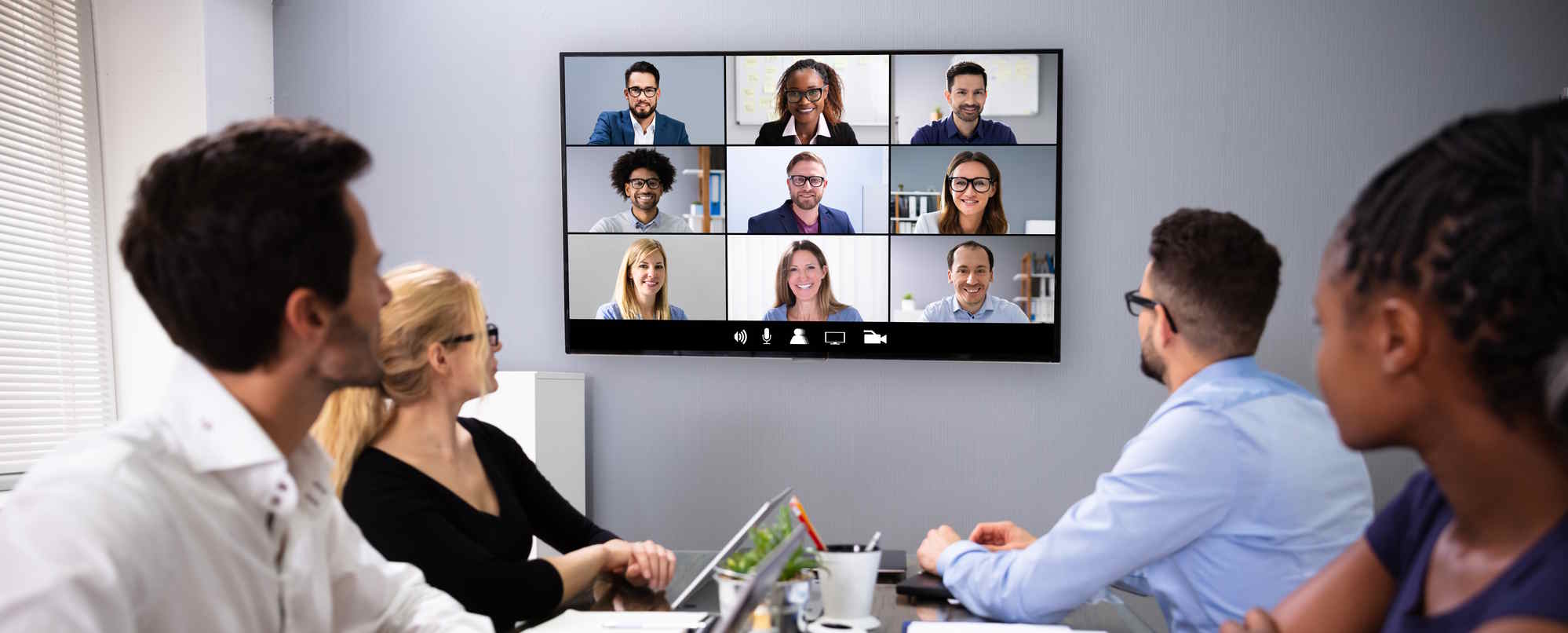 Hybrid meeting with four people at a conference table watching a 3×3 video call on a wall‑mounted screen.