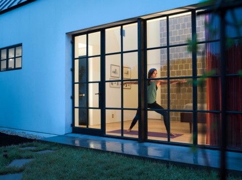 An exterior view into a house’s large windows. We see a woman doing yoga under warm lighting. 