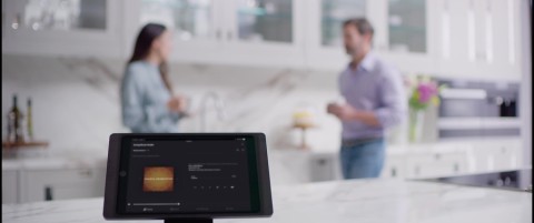 Touchscreen tablet in the foreground displaying music streaming. In the background, a man and woman chat in the kitchen. 