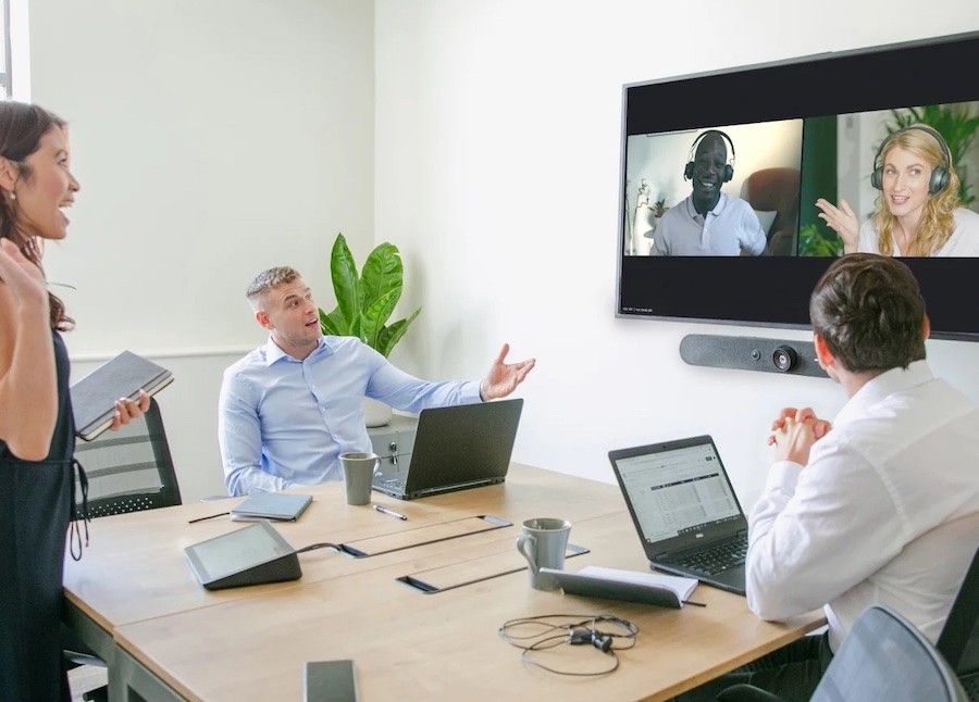 Three people in a small meeting room talking happily to two colleagues over a video call. On the table is a Logitech meeting room controller.