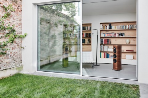A grassy lawn with a brick wall. A sliding door open to reveal a wooden Sonus Faber home audio speaker in a room with bookshelves. 
