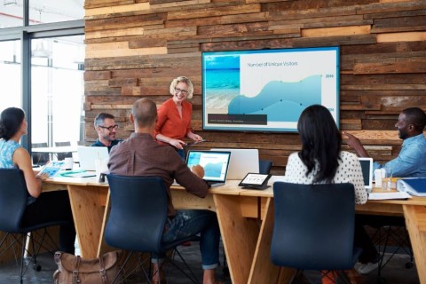 A huddle space with six participants with laptops or tablets, a display, and a control panel.