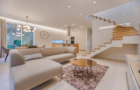 A brightly lit open-concept living room with a couch and stairs in the foreground and a dining room and kitchen in the background.