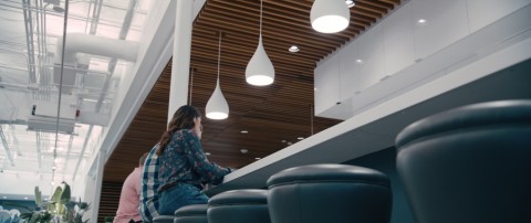 People sitting at a long table in an open concept office with commercial lighting fixtures hanging above.