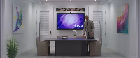 Man in a conference room with a video display on the wall.