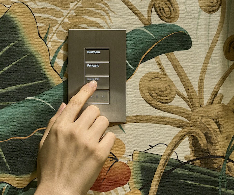 A hand touching a Lutron lighting keypad on floral wallpaper.