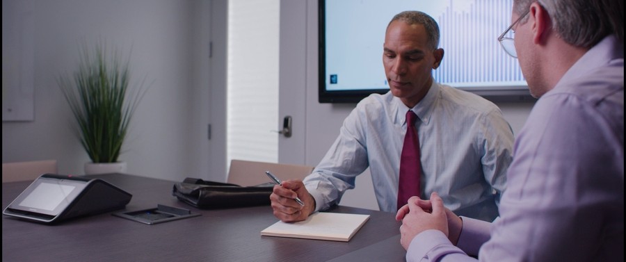 Man in a conference room with a tabletop controller and video display behind him. 