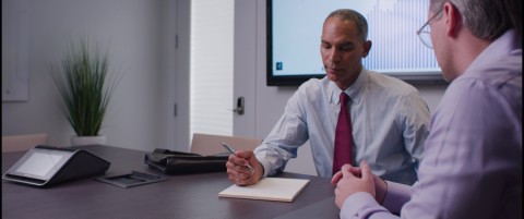 Man in a conference room with a tabletop controller and video display behind him. 