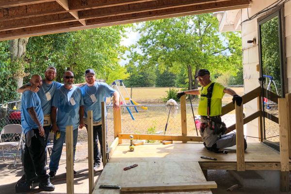 Five workers building a wooden porch, four in matching blue shirts posing and smiling on the left while another in a yellow safety vest works on the right, with lumber, tools, and a grassy yard with trees and a swing set in the background.