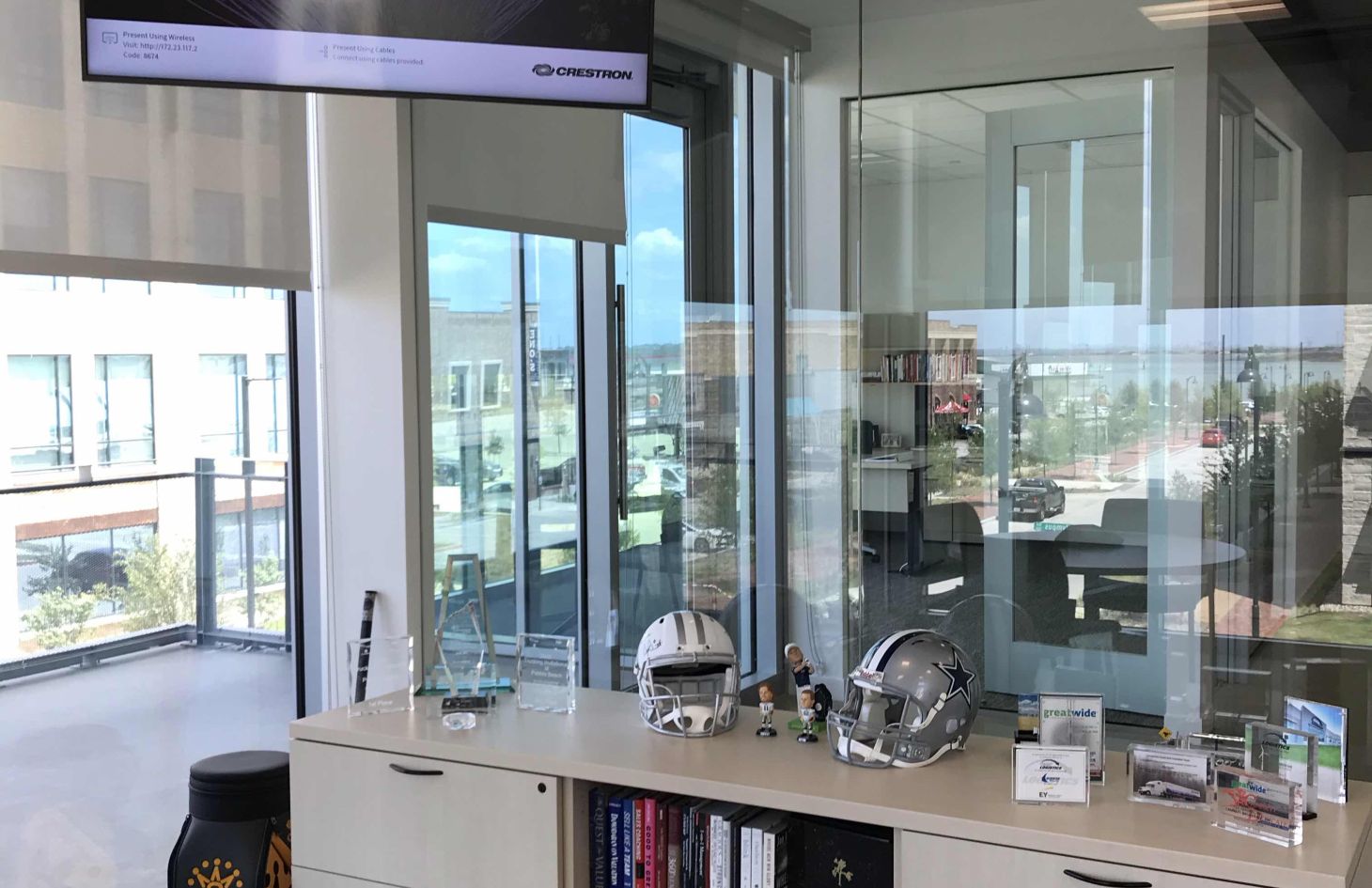 Modern corner apartment living and dining area with floor-to-ceiling windows and roller shades, a gray sectional sofa, round dining table with four chairs, and a large white column on light wood floors overlooking a city skyline.