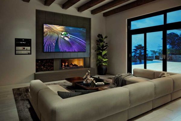 A modern living room with exposed ceiling beams, a large wall-mounted TV displaying a colorful image, and a built-in fireplace beneath. Floor-to-ceiling glass doors open to an outdoor view on the right.