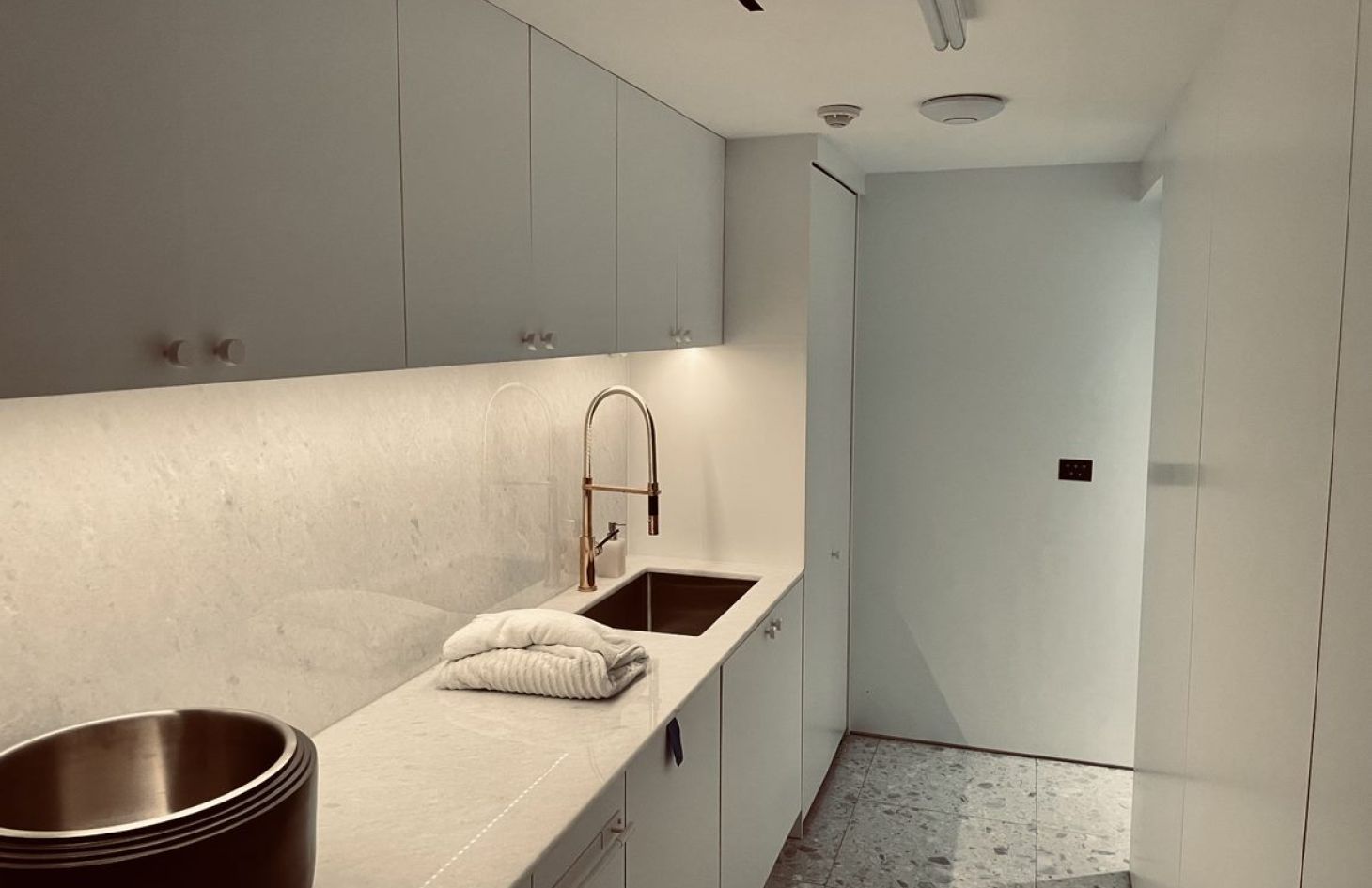 Minimalist galley-style kitchen with white cabinets, under-cabinet lighting, a stainless-steel sink and tall faucet, folded towels on the light countertop, and a metal bowl in the foreground.