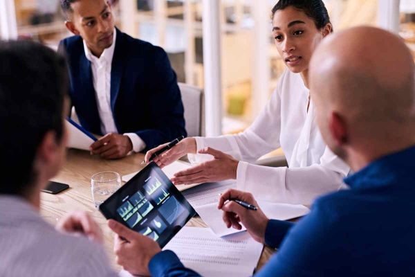 A professional team seated around a conference table, actively discussing business analytics and charts shown on a handheld tablet.