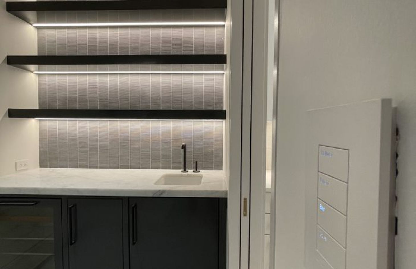 Modern kitchenette with dark lower cabinets, a small white sink, gray vertical tile backsplash, and three floating shelves lit by integrated LED strip lighting, with a wall-mounted touch control panel visible on the right.