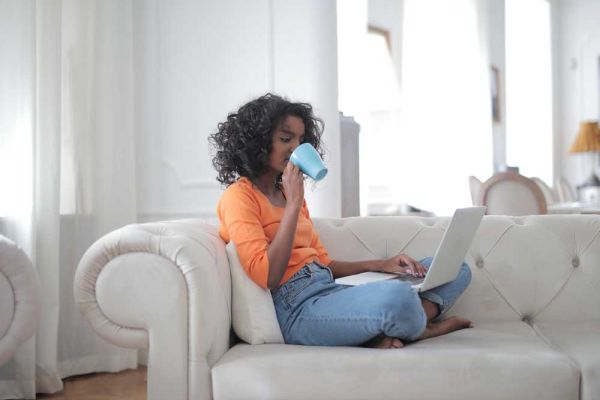 A woman in a bright orange top and jeans sits on a white sofa, sipping from a mug while using a laptop. Natural light filters into the minimalist living space.