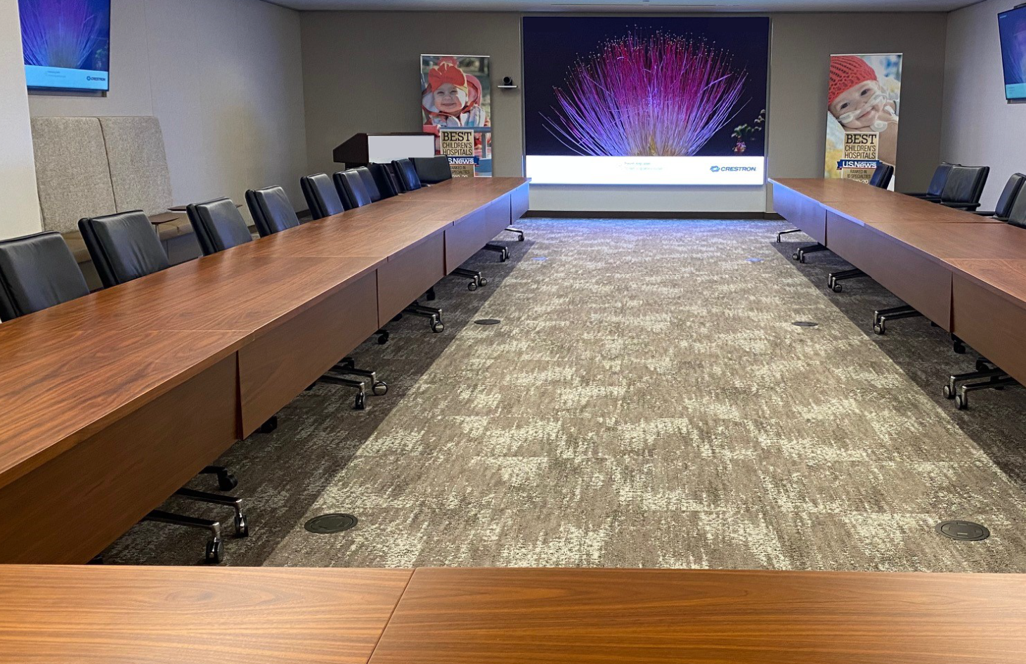 A long, rectangular conference room with a large presentation screen at the far end, flanked by posters featuring children&rsquo;s images. Rows of wooden tables and black chairs face the screen.