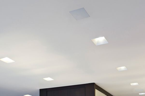 A perspective view of a clean, white ceiling featuring integrated square recessed lighting fixtures and a ceiling-mounted speaker or sensor.