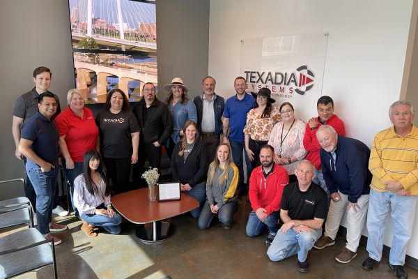 Group photo of about 18 Texadia Systems team members gathered in an office lobby, some standing and some seated around a small table with a framed certificate, with a large Texadia Systems logo sign and a city bridge photo on the wall behind them.