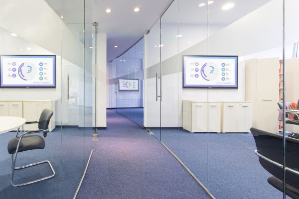 A modern, bright office corridor featuring glass partitions that reveal conference rooms equipped with digital displays showing business analytics and "Vertical Markets" data.
